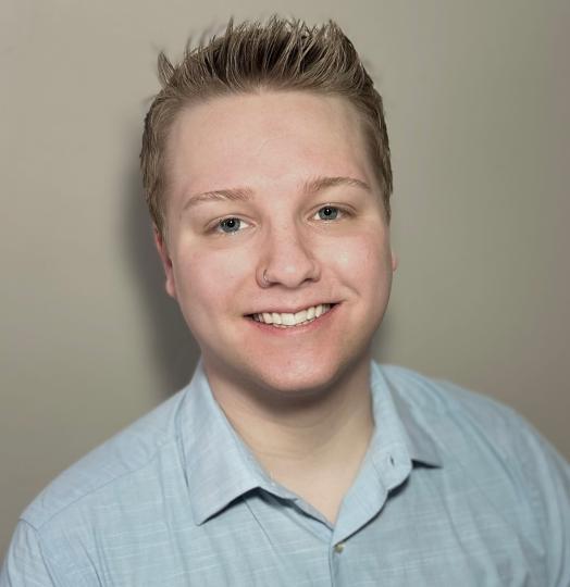 white male with crew cut hair cut, nose ring, and a light blue shirt with a cream colored background.
