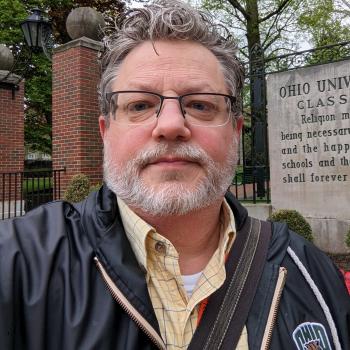 Picture of a man in yellow plaid dress shirt and Ohio University jacket standing. 