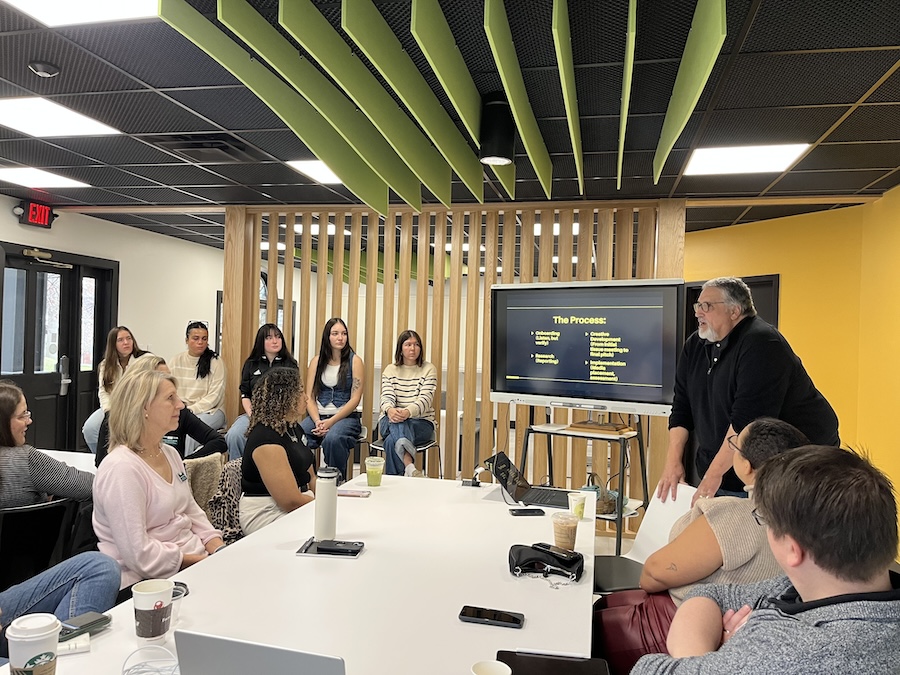 A man stands before a group of people in a conference room, presenting