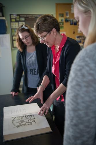 Two students stand with a professor examining a large book open in front of them