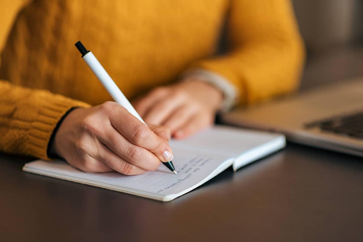 Close-up of hands writing in a notebook with a laptop open nearby