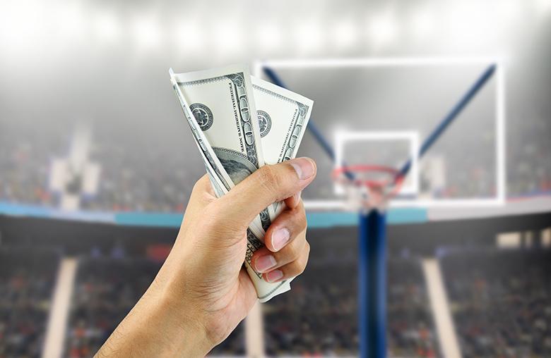 A hand holds money with a basketball court in the background.