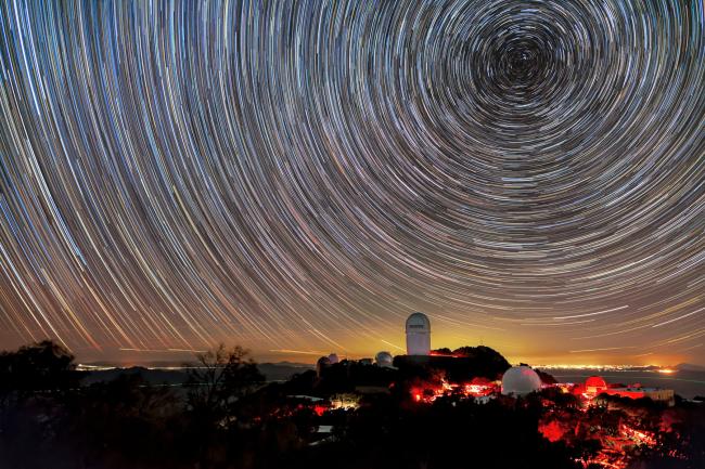  A long-exposure image captures circular star trails above telescope domes illuminated in red light on a dark mountain. A golden glow on the horizon marks distant city lights.