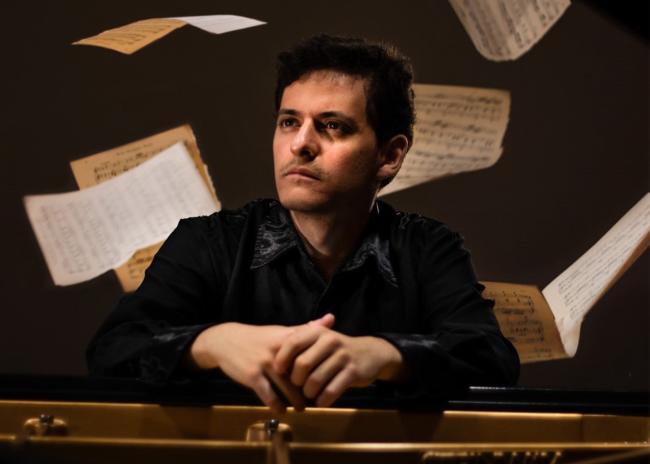Alejandro Orta, seated at the piano looking off to the side while sheets of music float behind him
