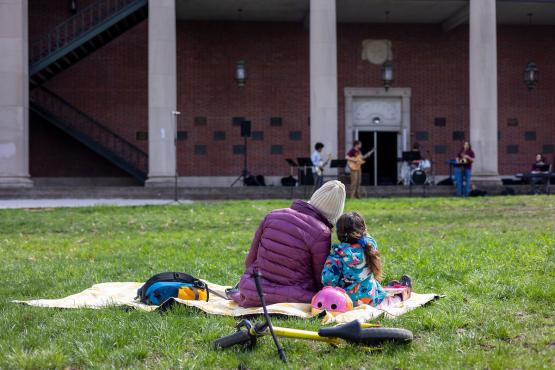 A parent and child sit on the College Green, with their bikes nearby, while listening to performers play music