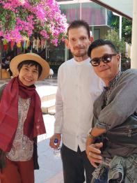 Three people standing in front of a pink blossom tree next to a builidng in Thialand