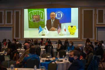 Chubu University Professor Nobuyuki Kawauchi giving an acceptance speech broadcasted on a screen while onlookers watch from dining tables