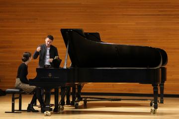 Two people on stage, one sitting at a piano, and the other standing