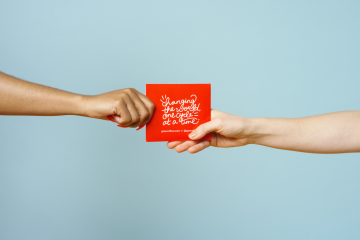An arm reaches from the left handing a period product in an orange box to an arm reaching from the right