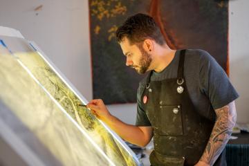 An artist stands at an illuminated easel, working on a print