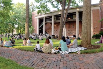 People sit on the grass near Memorial Auditorium, listening to an orchestra playing on College Green.