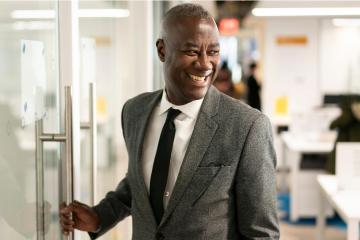 Byron Ward stands in a grey suit, smiling and looking back as he opens a door.