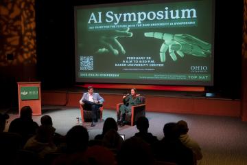 Two speakers sit in front of an audience in front of a large screen reading "AI Symposium" 