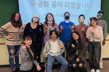A group poses in front of a classroom, smiling and making a gesture with their hands