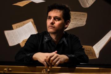 Alejandro Orta, seated at the piano looking off to the side while sheets of music float behind him
