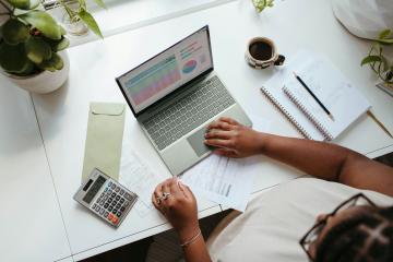 overhead view of a person typing on a laptop, surrounded by notebooks, a calculator, and a cup of coffee