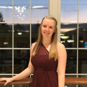 Lillie Clepper poses in front of a large window, with a hand placed on a wooden railing that rests behind. Lillie wears a formal, one-shoulder maroon dress.