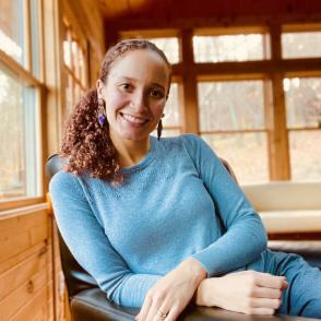 Paola García Morales sits leaning against the left arm of a leather chair, Paola García Morales smiles at the camera, wearing a light blue long sleeve shirt and diamond-shaped earrings.