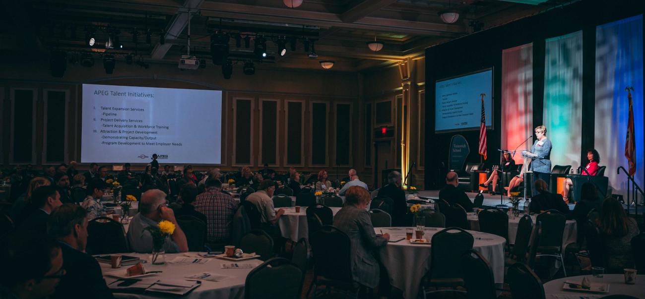 Large conference room with audience seated at tables and speaker on stage
