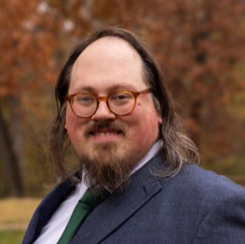 Paul Valentine Headshot. Man with brown hair and goatee, with read glasses, wearing a dark blue suit jacket and green tie. 