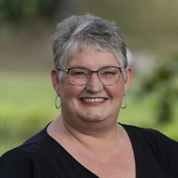 Carleen Dotson's profile picture Headshot of Carleen Dotson. Woman smiling at the camera with short gray hair, dark wire framed glasses, earrings and a dark v-necked top. The background of the photo is blurred outdoor scenes with grass and trees.