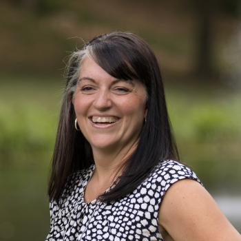 Erin Gibson's profile picture Headshot of Erin Gibson. Woman smiling towards camera with shoulder length dark hair, wearing a printed top. The background is outdoors with grass and trees behind her.