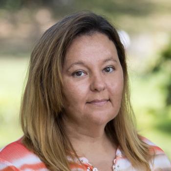 Teri Clapper's profile picture Headshot of Teri Clapper. A woman smiles at the camera, she is wearing a coral and white patterned shirt, with highlighted brown hair. The background of it image is unfocused landscaping of trees, shrubs and grass.