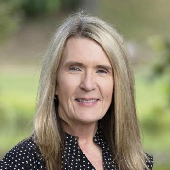 Headshot of Trenia Twyman. Woman smiles at the camera, she has long blonde hair and is wearing a black dress with white polka dots. The background of the image is of unfocused trees and landscaping.