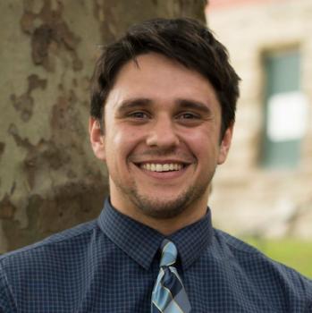 Headshot of Zarek Bell. Man with brunette hair smiles at the camera wearing a blue, collared shirt and patterned blue tie. In the background is a textured tree trunk and out of focus brick building. 