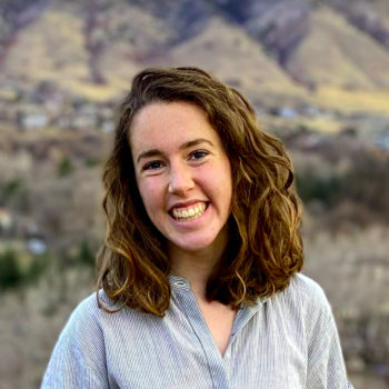 Portrait of Sarah Liederbach. Smiling woman looks at the camera, she has shoulder length brown hair and is wearing a light blue shirt. The backdrop is of a hilly landscape.